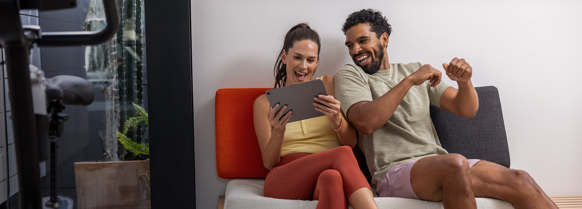 Man and woman sitting on a couch with a tablet, with gym equipment in the background