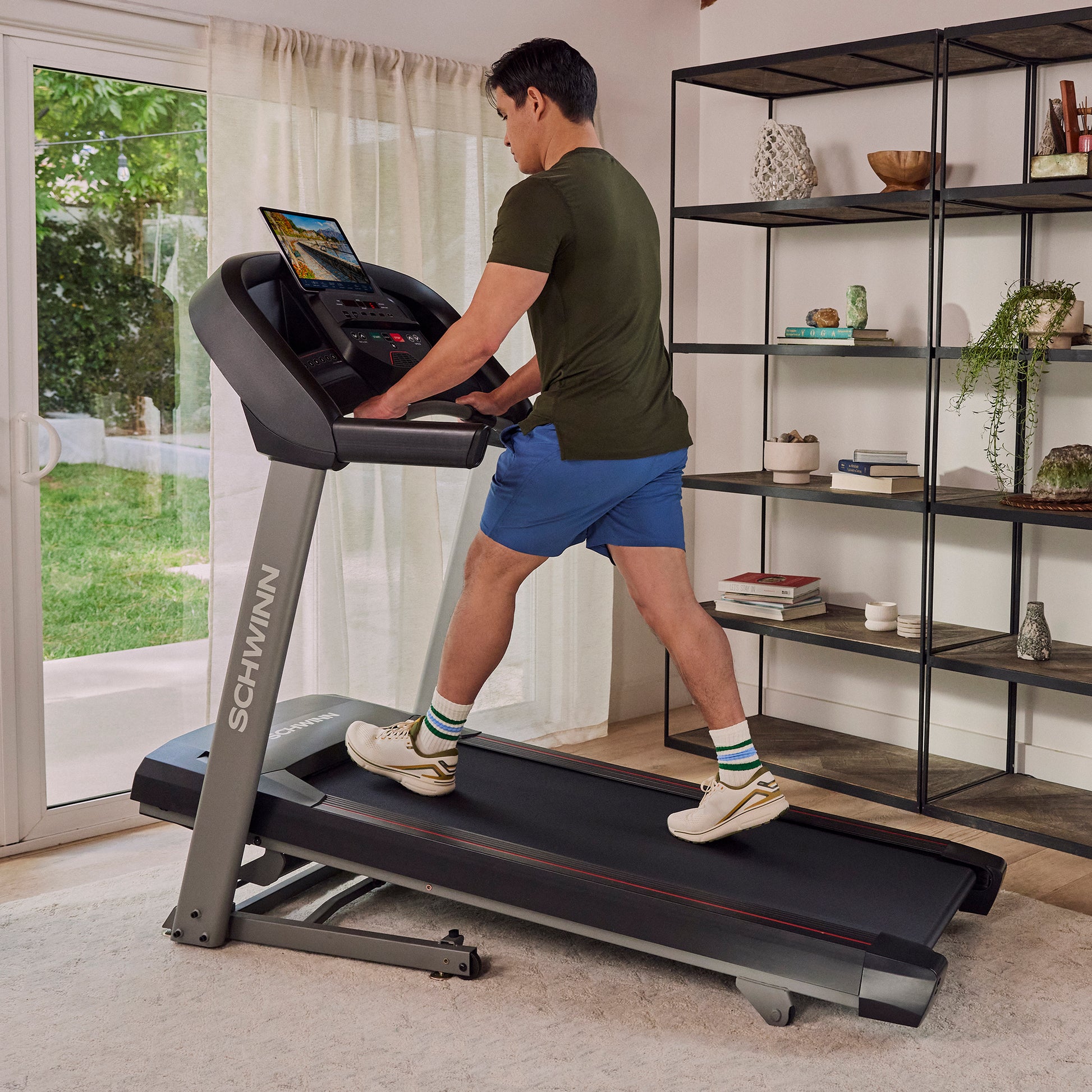 Man using a treadmill at an incline in a home setting with a bookshelf and window in the background